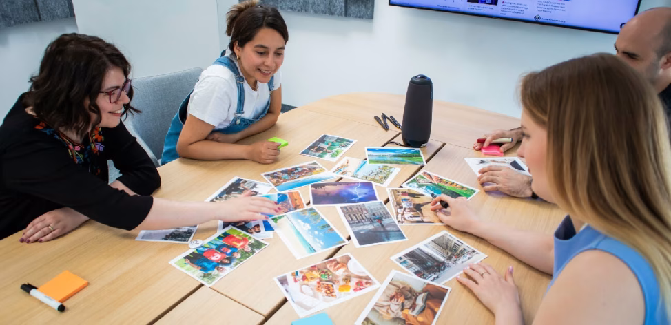 O echipă corporate diversă colaborează intens la o activitate experiențială de tip Board Games cu twist de Business sau o etapă dintr-o simulare de business complexă, organizată direct în sala lor de ședințe de Yes Academy, ca parte a unui program de team building la birou. Participanții analizează fotografii de training pentru a lua decizii strategice.