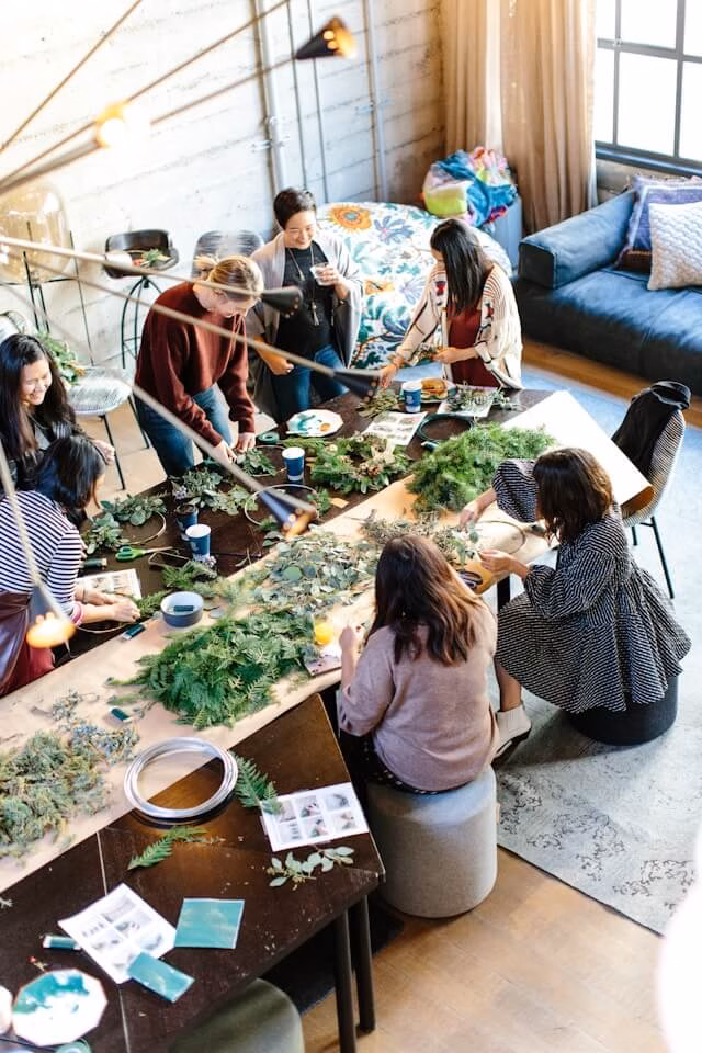 Un grup de angajați participă la un workshop creativ, realizând decorațiuni din plante în jurul unei mese mari, chiar la sediul companiei. Imaginea ilustrează o activitate de team building la birou relaxantă, organizată într-un spațiu de lucru modern, eliminând costurile de deplasare.